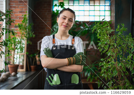Running of own business. Young woman florist wearing apron in botanical store with green plants. Happy small business owner working at flower shop standing surrounded by plants. Small business Running of own business. Young woman florist wearing apron in botanical store with green plants. Happy small business owner working at flower shop standing surrounded by plants. Small business 116629059