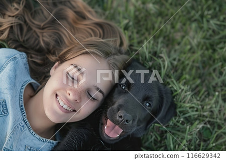 Portrait of kid playing with dog on a green grass yard. Child teen with a pet doggy in nature. Beautiful girl lying on lawn with puppy. Top view. Smiling happy owner girl cuddle with her best friend 116629342