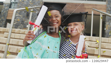 Image of gold confetti over happy diverse schoolgirls with diplomas and mortar boards embracing 116629447