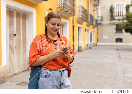 Happy young woman in sunglasses with mobile standing near yellow wall in old town. Holidays and tourism concept. Female tourist searching direction on smart phone while traveling abroad in summer Happy young woman in sunglasses with mobile standing near yellow wall in old town. Holidays and tourism concept. Female tourist searching direction on smart phone while traveling abroad in summer 116629486