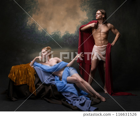 Classical-inspired portrait, with smiling woman draped in blue fabric, while standing man, wrapped in red, strikes confident pose. 116631526