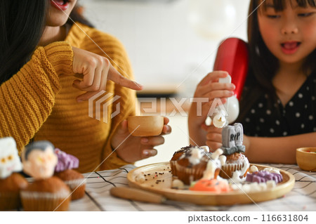 Beautiful young mother taking a picture of cute daughter with mobile phone decorating spooky cupcakes in kitchen 116631804