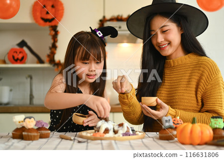 Beautiful young mother taking a picture of cute daughter with mobile phone decorating spooky cupcakes in kitchen 116631806