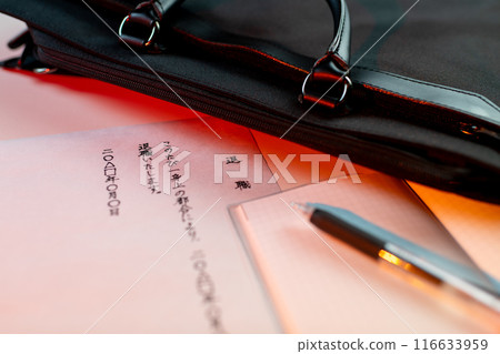 A handwritten resignation letter under dim red lighting, a gel-ink black pen on a notepad, and part of a business bag A handwritten resignation letter under dim red lighting, a gel-ink black pen on a notepad, and part of a business bag 116633959