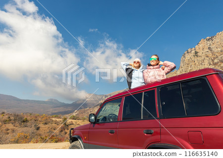 Scenic view two young adult woman friends enjoy having fun sit on car roof at adventure suv offroad mountain trip against blue sky on sunny autumn day. Female party nature trial journey concept Scenic view two young adult woman friends enjoy having fun sit on car roof at adventure suv offroad mountain trip against blue sky on sunny autumn day. Female party nature trial journey concept 116635140