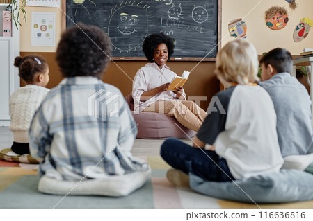 Young female teacher of Black ethnicity in bean bag chair reading book together with children sharing thoughts about story while sitting on floor in classroom Young female teacher of Black ethnicity in bean bag chair reading book together with children sharing thoughts about story while sitting on floor in classroom 116636816