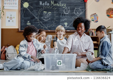 Multiethnic group of children and female teacher in ecology club sorting waste putting plastic bottles in recycling bin in classroom 116636824