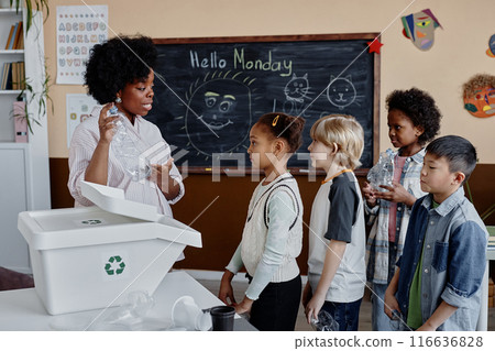 Young teacher of Black ethnicity at primary school explaining most common plastic types difference to children holding recyclable bottles in classroom with blackboard Young teacher of Black ethnicity at primary school explaining most common plastic types difference to children holding recyclable bottles in classroom with blackboard 116636828