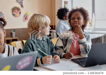 Multiethnic classmates working in pair with enthusiasm while using laptop sitting at desk at school Multiethnic classmates working in pair with enthusiasm while using laptop sitting at desk at school 116636878