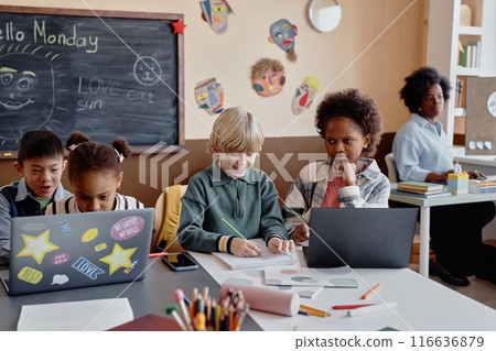 Shot of multiethnic group of children studying in pairs at large desk using laptops in primary school classroom decorated with blackboard and kids crafts Shot of multiethnic group of children studying in pairs at large desk using laptops in primary school classroom decorated with blackboard and kids crafts 116636879