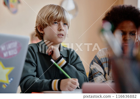 Portrait shot of studious blond haired boy in elementary school writing notes with pencil listening to teacher during lesson at school 116636882