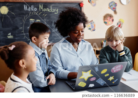 Happy female teacher of Black ethnicity using laptop explaining new theme to diverse group of kids around her in elective class 116636889