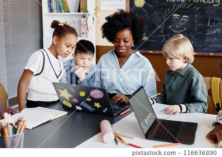 Diverse group of primary learners around female teacher of Black ethnicity explaining new information using laptop sitting at desk during elective lesson Diverse group of primary learners around female teacher of Black ethnicity explaining new information using laptop sitting at desk during elective lesson 116636890