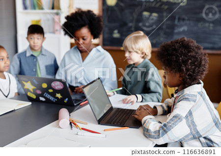 Side view of curly studious girl in primary school using laptop in smart classroom while female teacher explaining new topic with help of computer, copy space Side view of curly studious girl in primary school using laptop in smart classroom while female teacher explaining new topic with help of computer, copy space 116636891