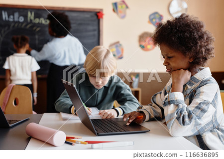 Side view of focused Black girl using laptop in diverse primary school while learning diligently at desk in class, copy space 116636895