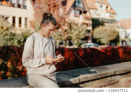 Young Fashionable Teenage Girl With Smartphone In Europian Park In Autumn Sitting At Smiling. Trendy Young Woman In Fall In Park Texting. Retouched, Vibrant Colors. Beautiful Blonde Teenage Girl 116637511