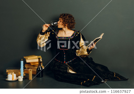 Young adult woman dressed in a medieval dress sitting near ancient books and candles with bottle of red wine and drinking a glass of wine 116637927