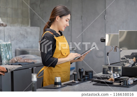 Young Female Barista in Yellow Apron Using Tablet in Modern Coffee Shop with Professional Equipment 116638016