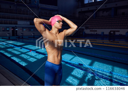 Young man with athletic body, swimmer wearing goggles and pink cap, posing against blurred background of pool. 116638046