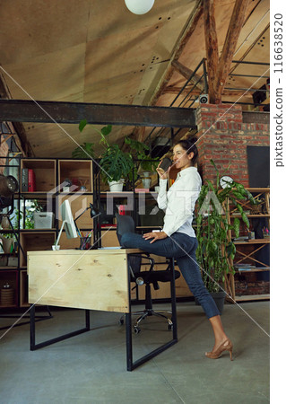 Promoting healthy and active lifestyle in modern office setting. Woman standing at her desk, stretching and drinking coffee Promoting healthy and active lifestyle in modern office setting. Woman standing at her desk, stretching and drinking coffee 116638520