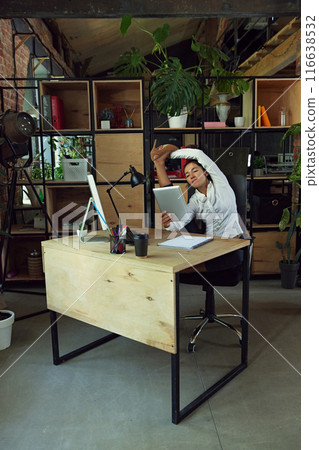 Wo,man. office worker sitting at desk in stretching position, working on tablet against modern office settings 116638532