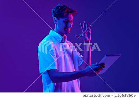 Young man in white polo shirt using tablet for online video call, waving for greetings against gradient blue purple background in neon light 116638609