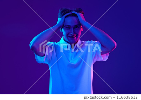 Portrait of young emotional man in white polo shirt, holding hand son head with shocked and excited face against gradient blue purple background in neon light 116638612
