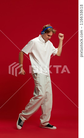 Full-length of young handsome man in white polo shirt wearing blue headphones, listening to music and dancing against red studio background 116638618