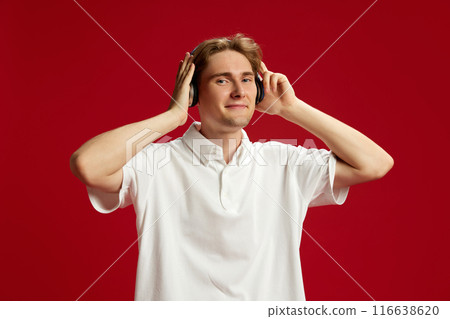 Smiling young handsome man in white polo shirt wearing blue headphones and listening to music against red studio background 116638620