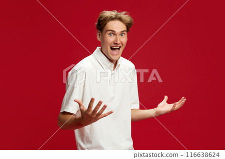 Young man in white polo shirt spreading hands in positive shock and excitement against red studio background. Sales, success, achievement 116638624