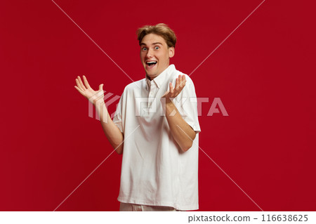 Young man in white polo shirt spreading hands in positive shock and excitement against red studio background. Sales, news 116638625