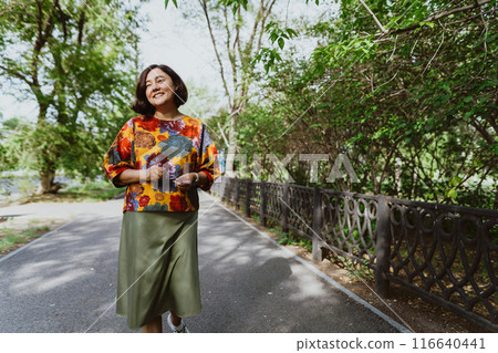 Asian woman with a joyful, open smile enjoys the tranquility of a summer day while walking along the sidewalk of the park Smiling middle aged woman walking, relaxing in park and radiating happiness Asian woman with a joyful, open smile enjoys the tranquility of a summer day while walking along the sidewalk of the park Smiling middle aged woman walking, relaxing in park and radiating happiness 116640441