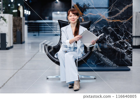 Woman in a light blue suit sitting in a black chair holding a white book in a modern showroom 116640805