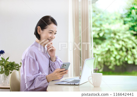 A middle-aged woman looking at a computer while using her smartphone in the living room 116642441
