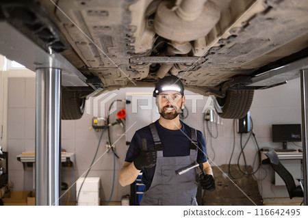 Mechanic giving thumbs up under raised car in auto repair shop 116642495