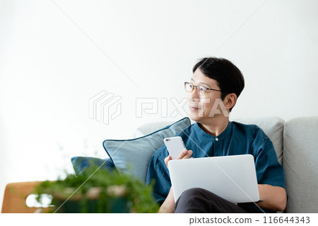 A middle-aged man operating a computer and smartphone on the sofa 116644343