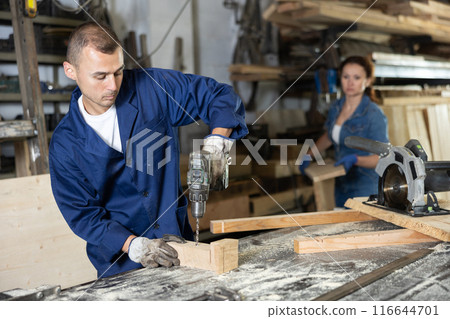 Male employee of carpentry workshop uses drill to make holes in wooden billet 116644701