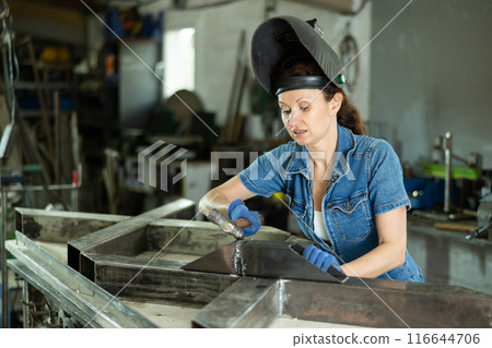 Portrait of a female welder standing with a welding semi-automatic machine and a safety helmet in metal machining workshop 116644706