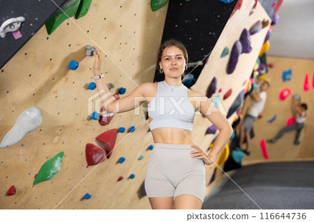 Positive slender girl in sportswear is standing near climbing wall in gym 116644736