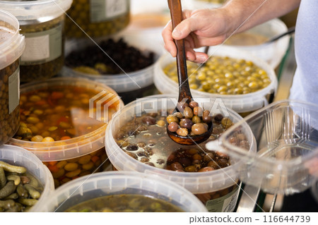 Mens hands hold ladle and place pickled black olives with stone in disposable plastic container Mens hands hold ladle and place pickled black olives with stone in disposable plastic container 116644739