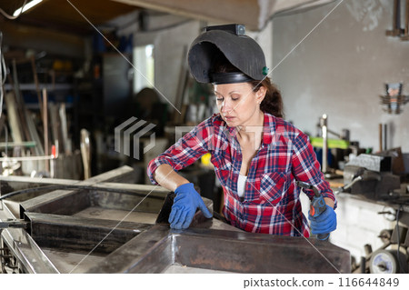 Portrait of a female welder standing with a welding semi-automatic machine and a safety helmet in metal machining workshop 116644849