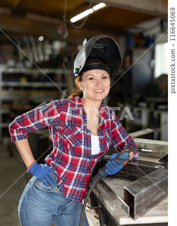 Portrait of a female welder standing with a welding semi-automatic machine and a safety helmet in metal machining workshop Portrait of a female welder standing with a welding semi-automatic machine and a safety helmet in metal machining workshop 116645069
