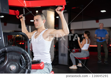 Portrait of man doing triceps curls in exercise machine during strength training in gym 116645291