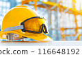 A yellow hard hat and safety goggles sit on a white surface, with a blurred construction site background. 116648192