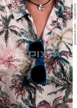 Close-up of blue sunglasses hanging from the Hawaiian shirt of a man wearing a necklace. 116656019
