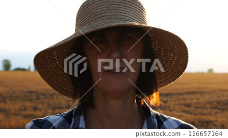 Serious female farmer in straw hat looking into camera against the blurred background of wheat field. Portrait of adult agronomist standing on barley meadow at sunset. Concept of agricultural business 116657164