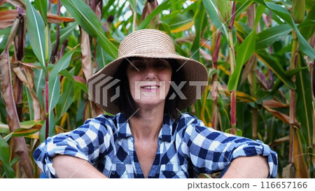 Happy smiling female farmer looks into camera sitting near corn field. Portrait of adult tired agronomist in straw hat with maize meadow at background. Concept of agricultural business. Close up 116657166