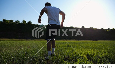 Sportsman juggling soccer ball on stadium at sunny day. Young man kicking ball at green field. Professional footballer practicing tricks at meadow with sunlight at background. Freestyle football. 116657192