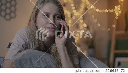 Pretty young lady chatting on mobile phone discussing news smiling lying in bed in illuminated bedroom in evening. People and cellphone communication concept. 116657472