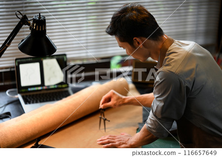 Close up of an artisan hands meticulously measuring a piece of leather using a pair of dividers in workshop 116657649